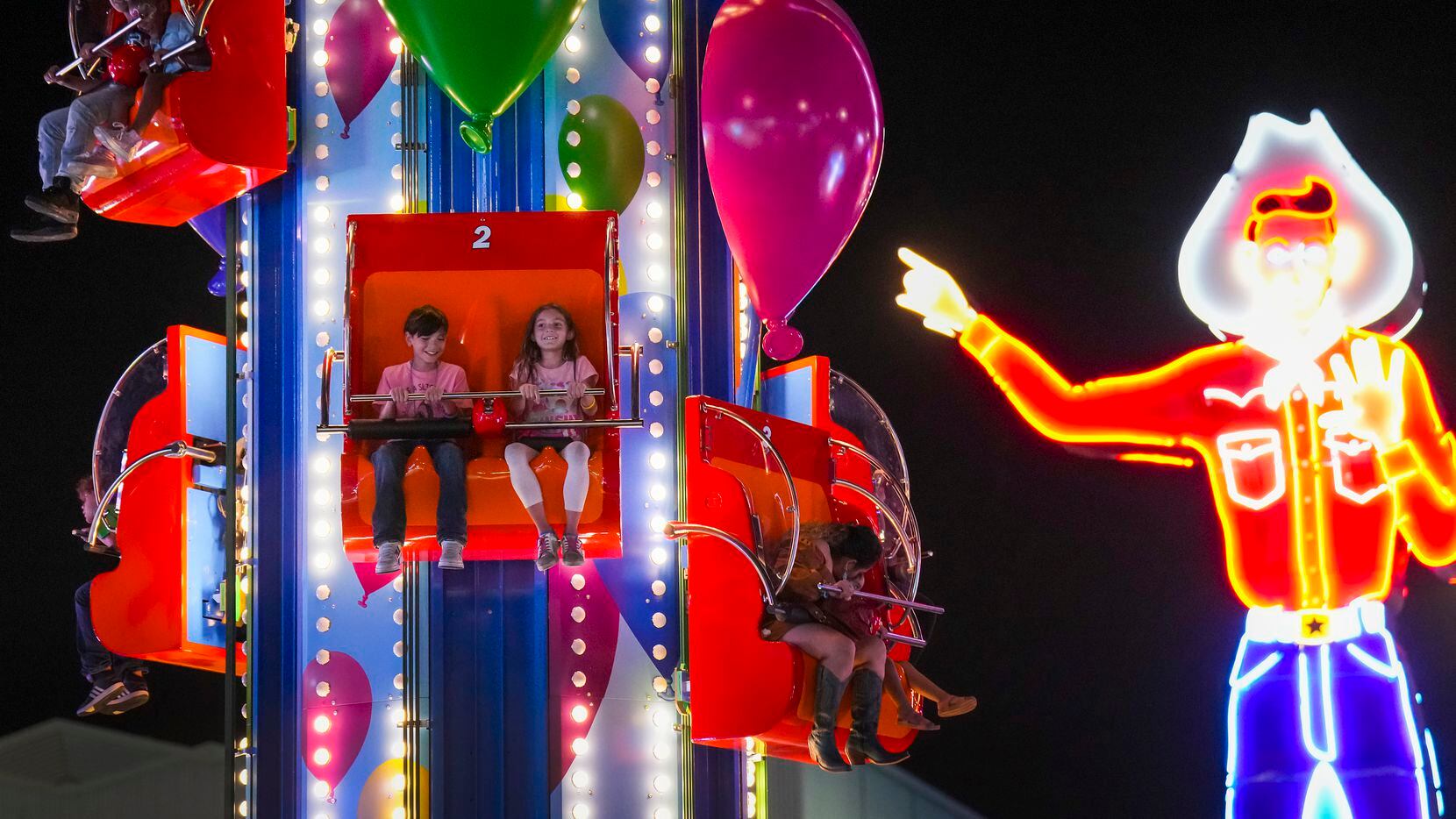 Neon Big Tex points toward youngsters on a ride on the midway on opening night at the State...