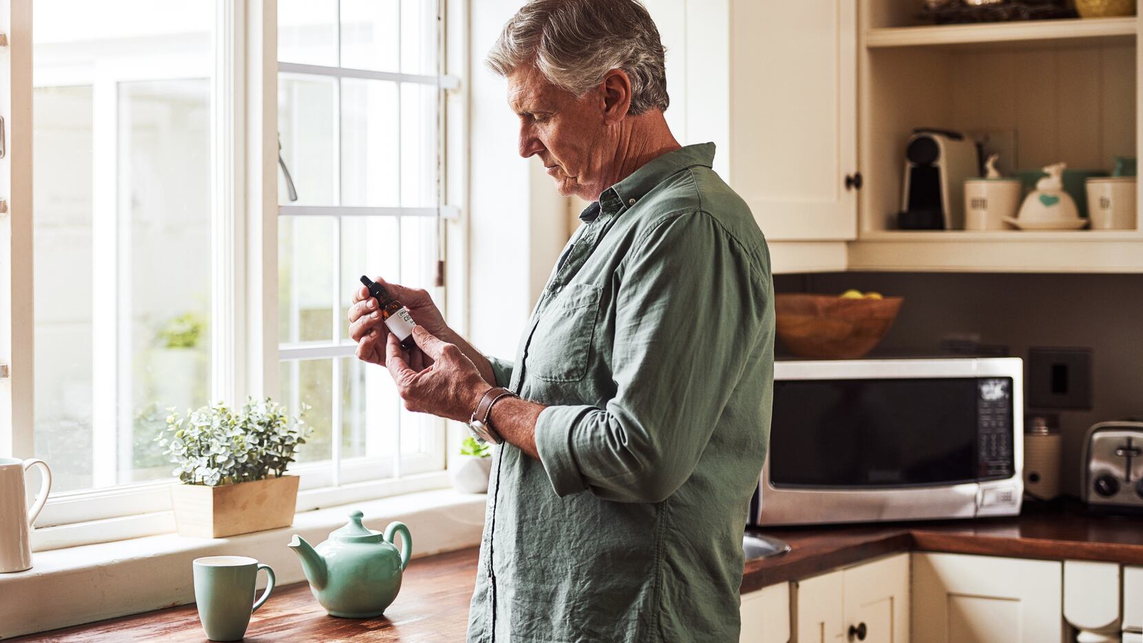 A older man examines a bottle of CBD oil at his kitchen counter, with a teapot and cup in...