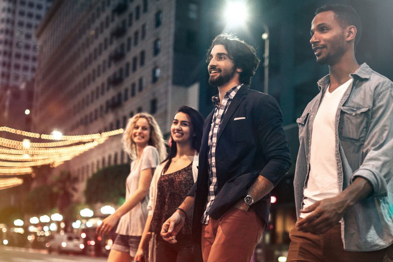 A group of multi-ethnic young adults explore and walk around downtown Los Angeles, California on a summer evening, checking out the city night life.  They walk casually down a city street lined with cars and tall buildings, talking and enjoying the night.  Horizontal image.