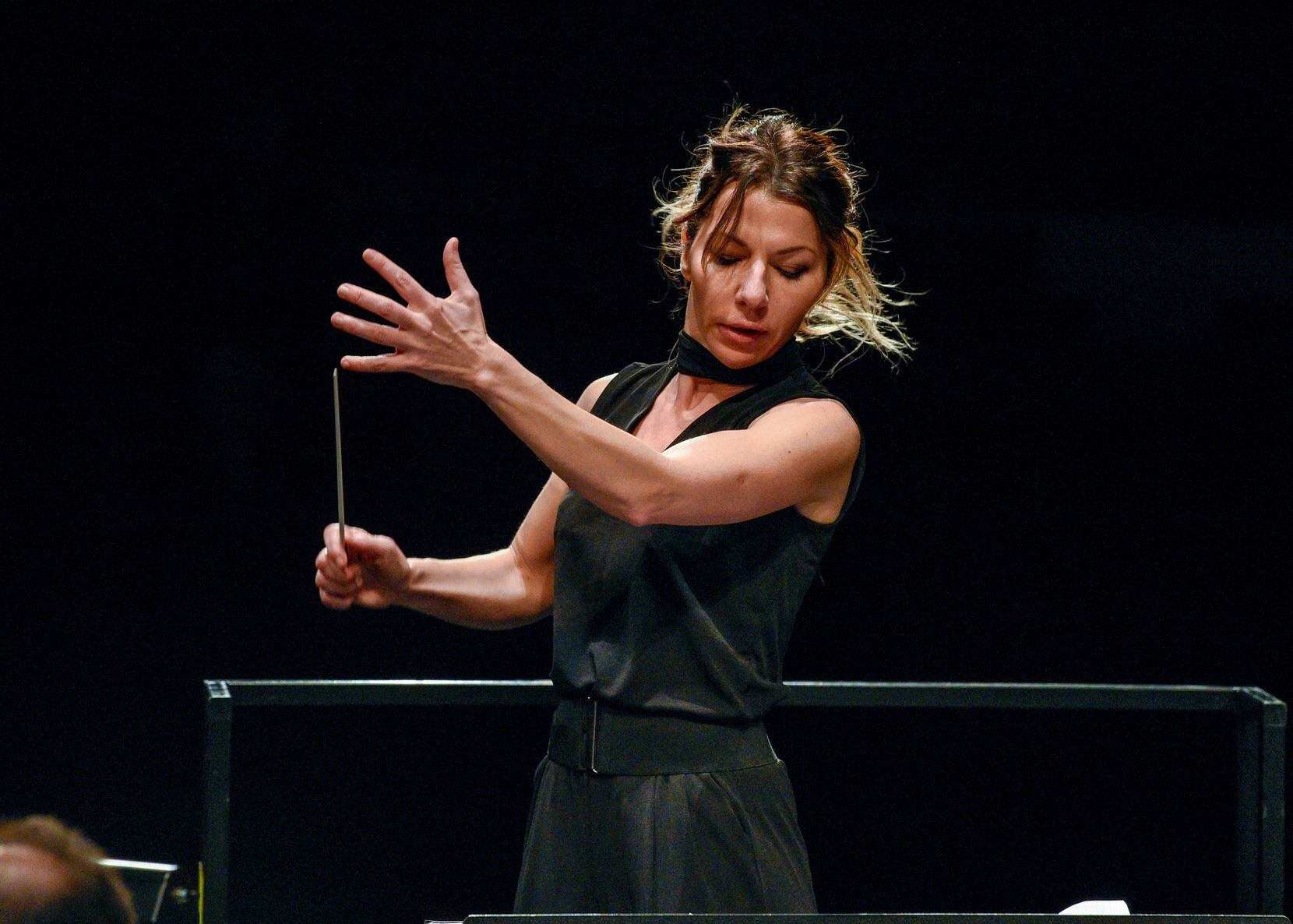 A blonde woman conductor holds a baton while conducting an orchestra.