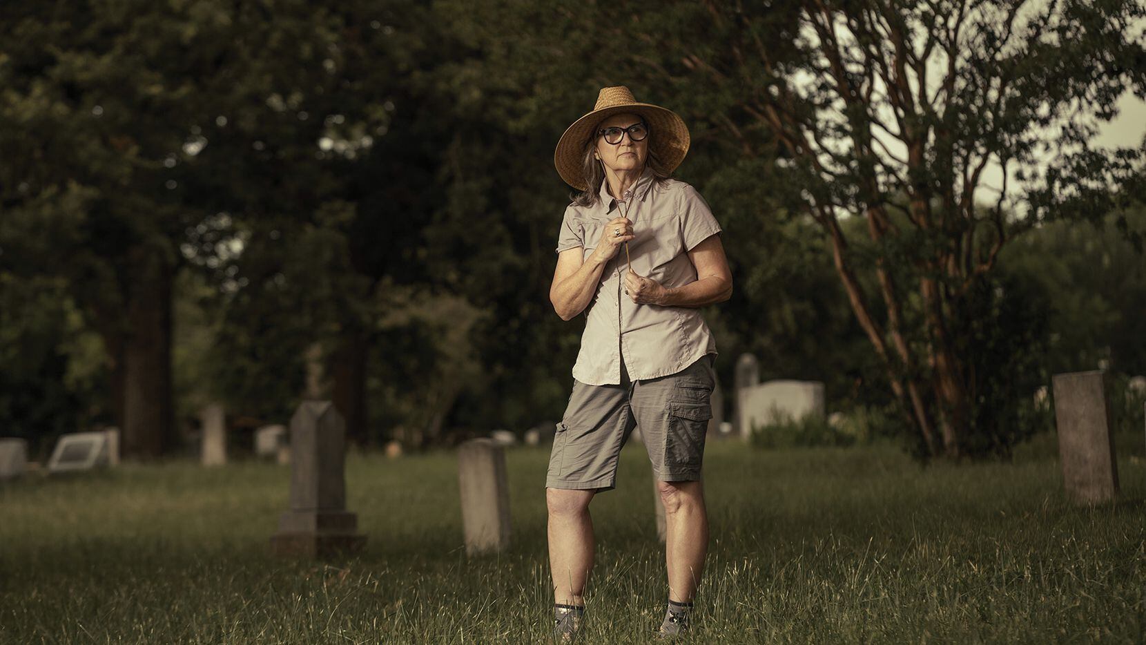 woman in work clothes and wide-brimmed hat surveys a cemetery