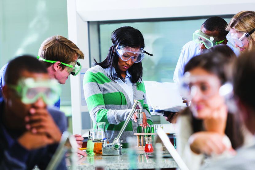 Multi-ethnic group of students with instructor in chemistry lab, wearing safety goggles,...