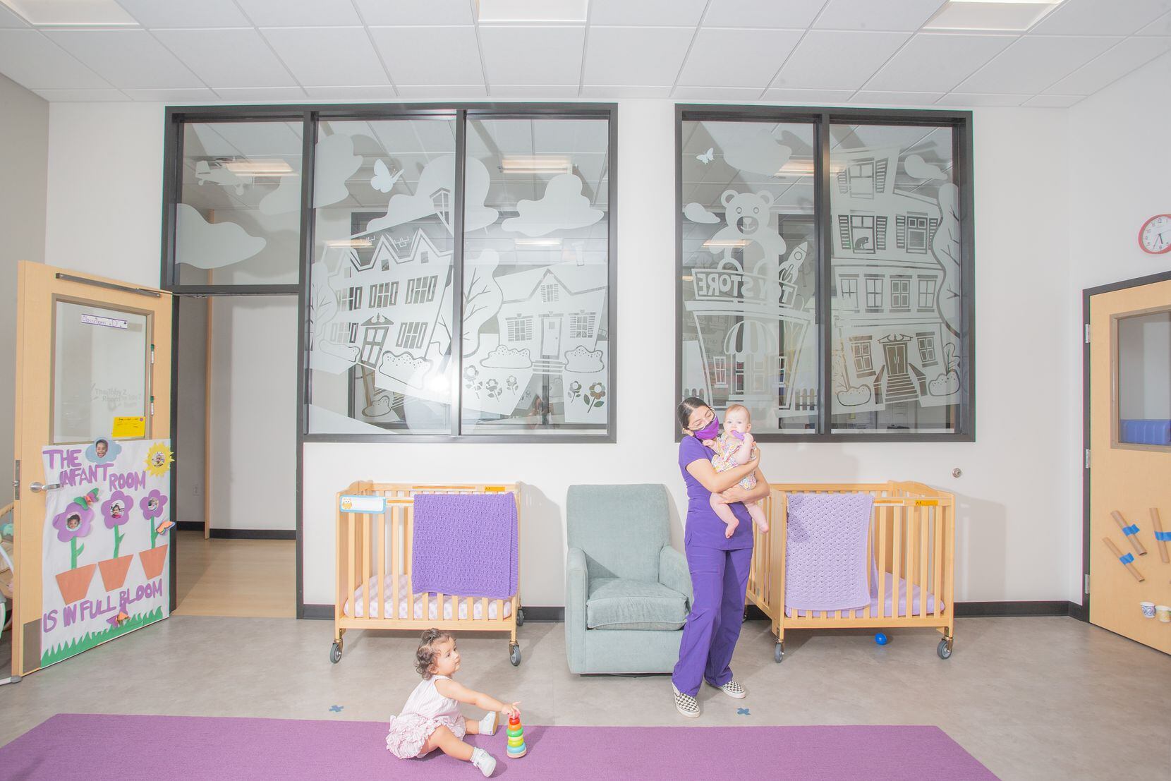 A nurse in purple scrubs holds a baby while a toddler plays on the floor in day care center.