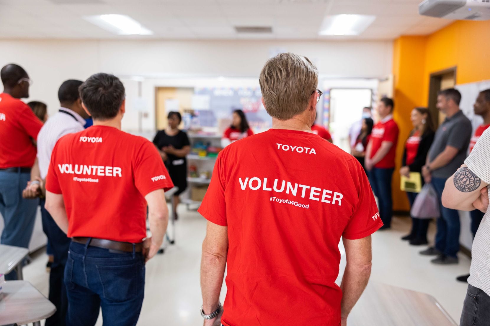 Several volunteers in red T-shirts stand in a classroom with other people.