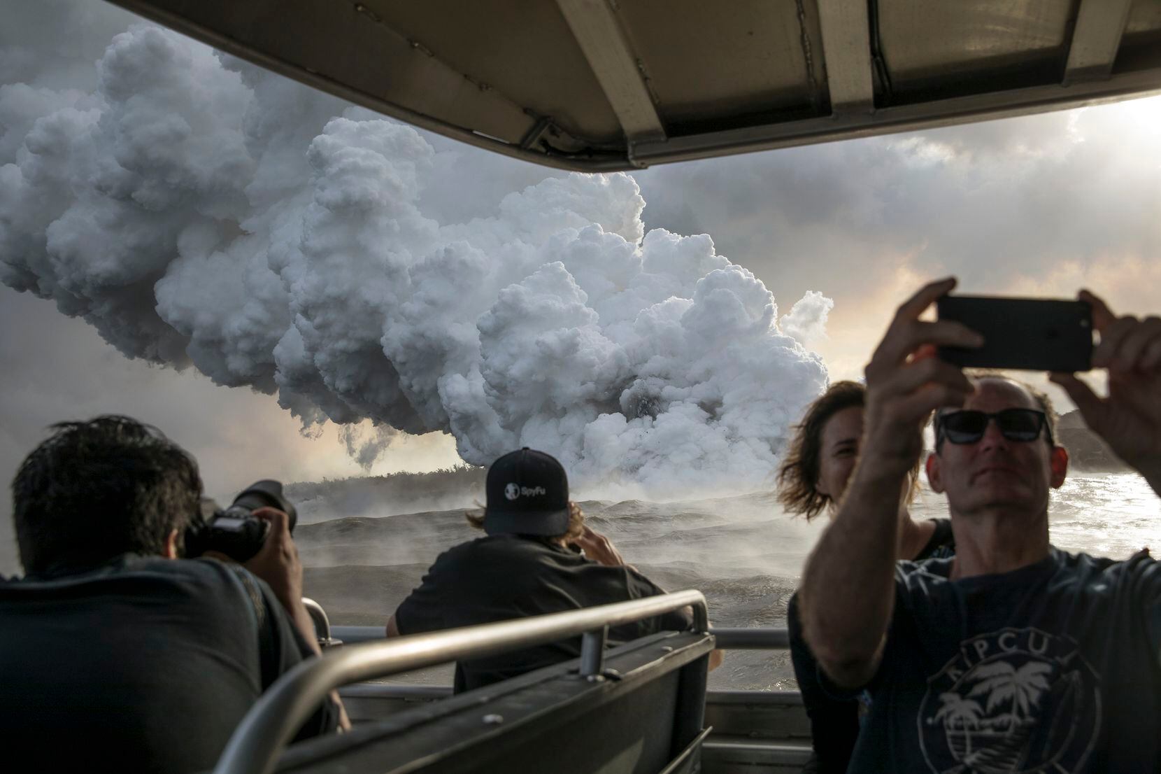 Passengers on a tour boat take photos of a plume of volcanic gases and steam rising as lava...