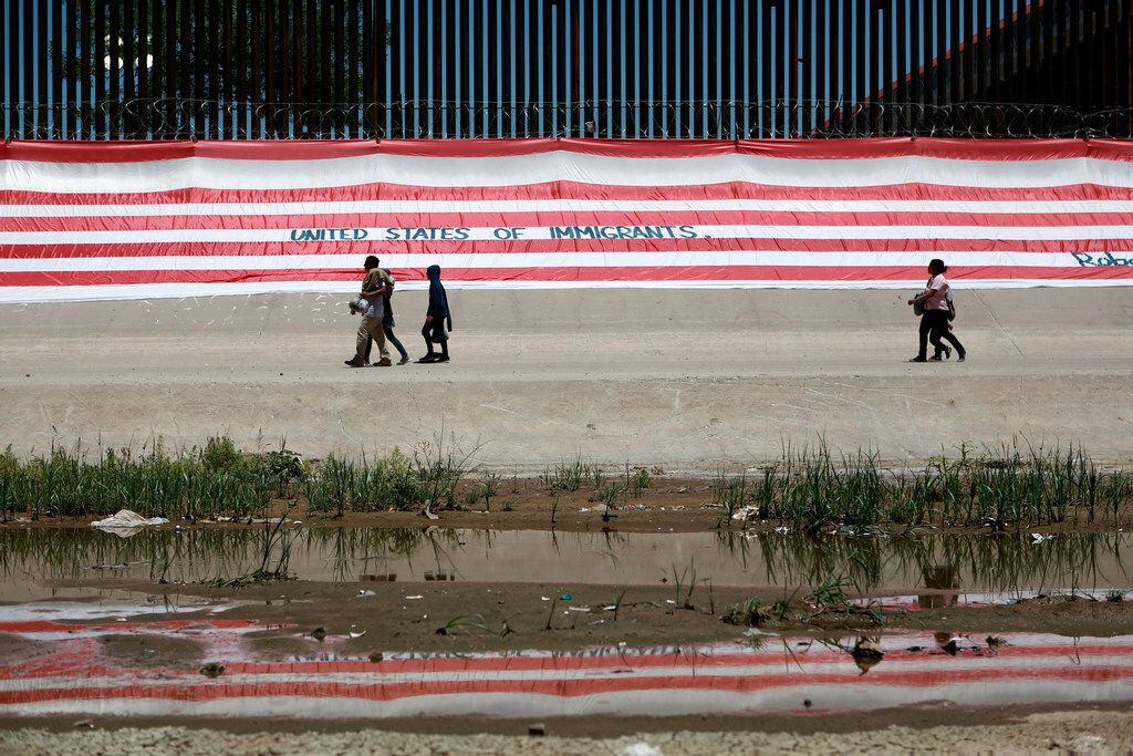 In this June 7, 2019 photo, migrants walk past a large U.S. flag blanketing a portion of the...
