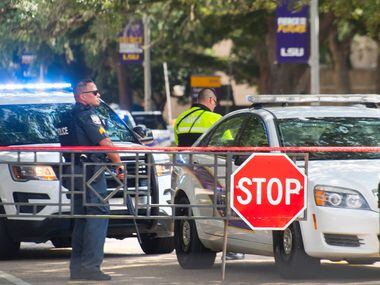 Law enforcement personnel stand with weapons near two LSU police vehicles parked between the...
