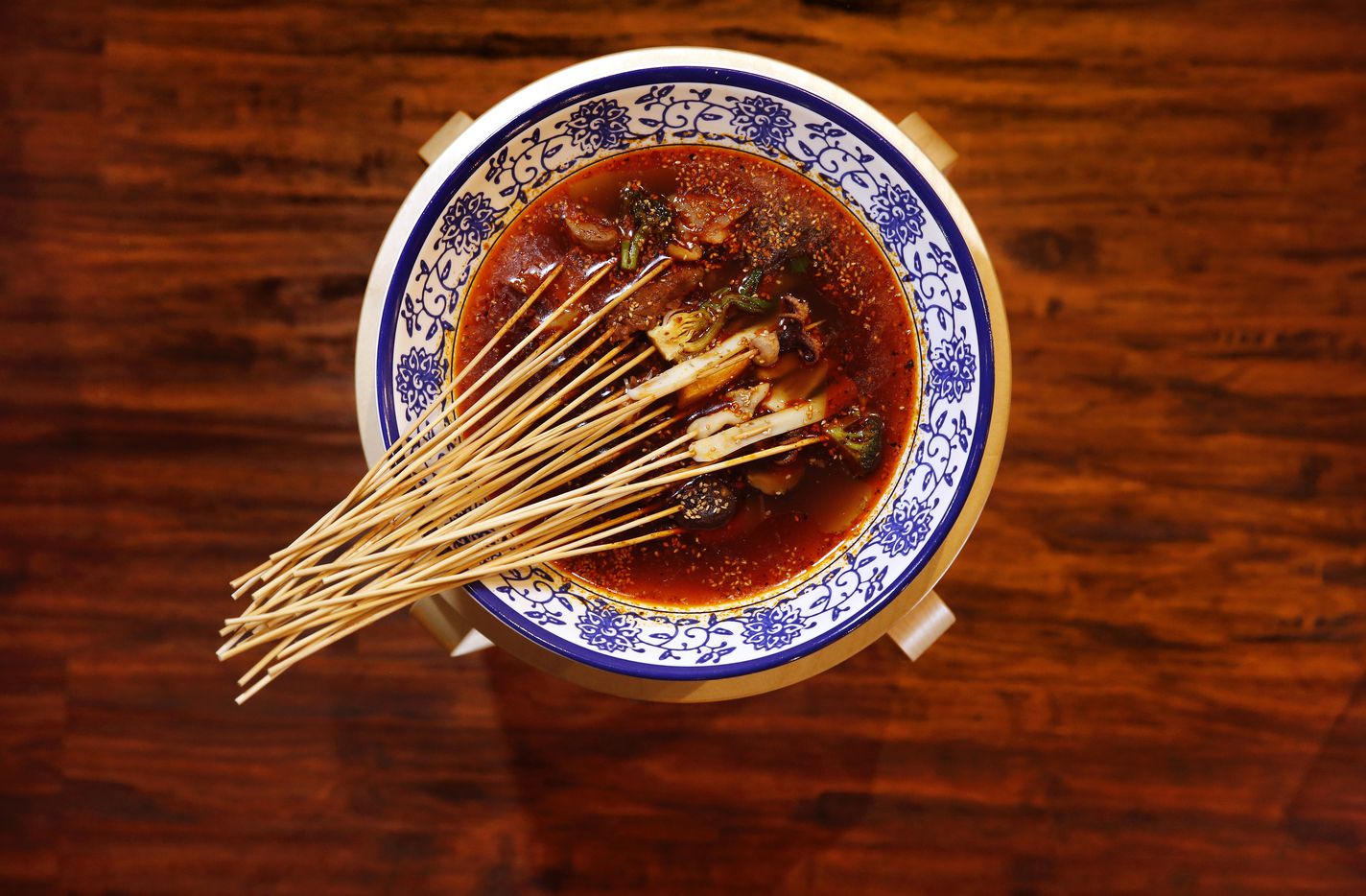 Skewered meats and seafood are served in a large bowl of spicy chicken broth with Sichuan...
