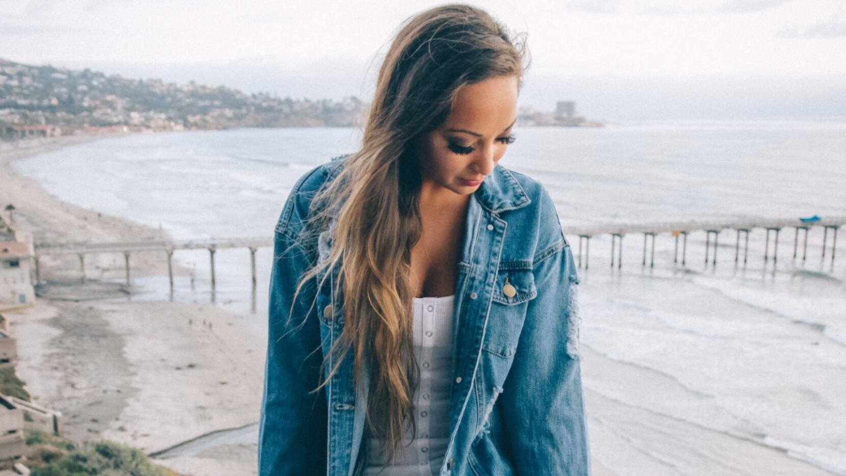 Sarah P Antonella wears a denim jacket while looking down while the beach is in the background