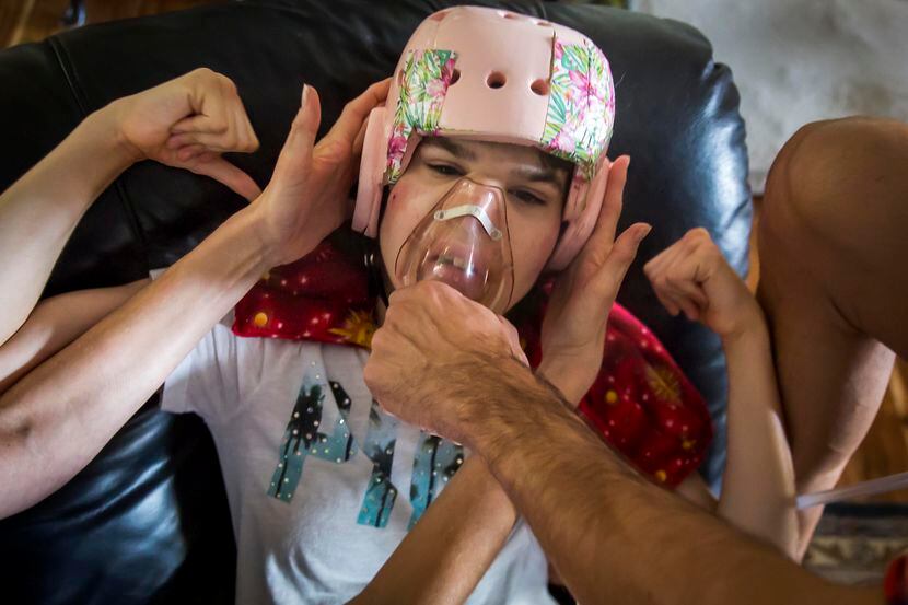 Kara Zartler punches herself in the head as her father Mark places a medical mask over her...