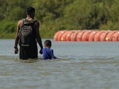 Migrants walk past large buoys being used as a floating border barrier on the Rio Grande...