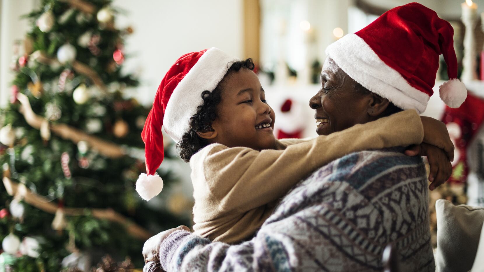 A parent and child hug while wearing Santa hats with a Christmas tree in the background.