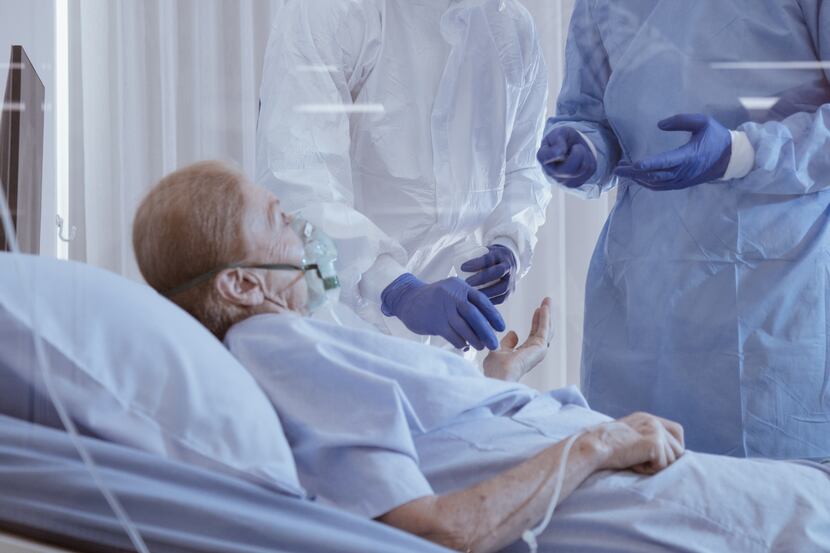 A woman lays in a hospital bed wearing a breathing mask while doctors stand over her.