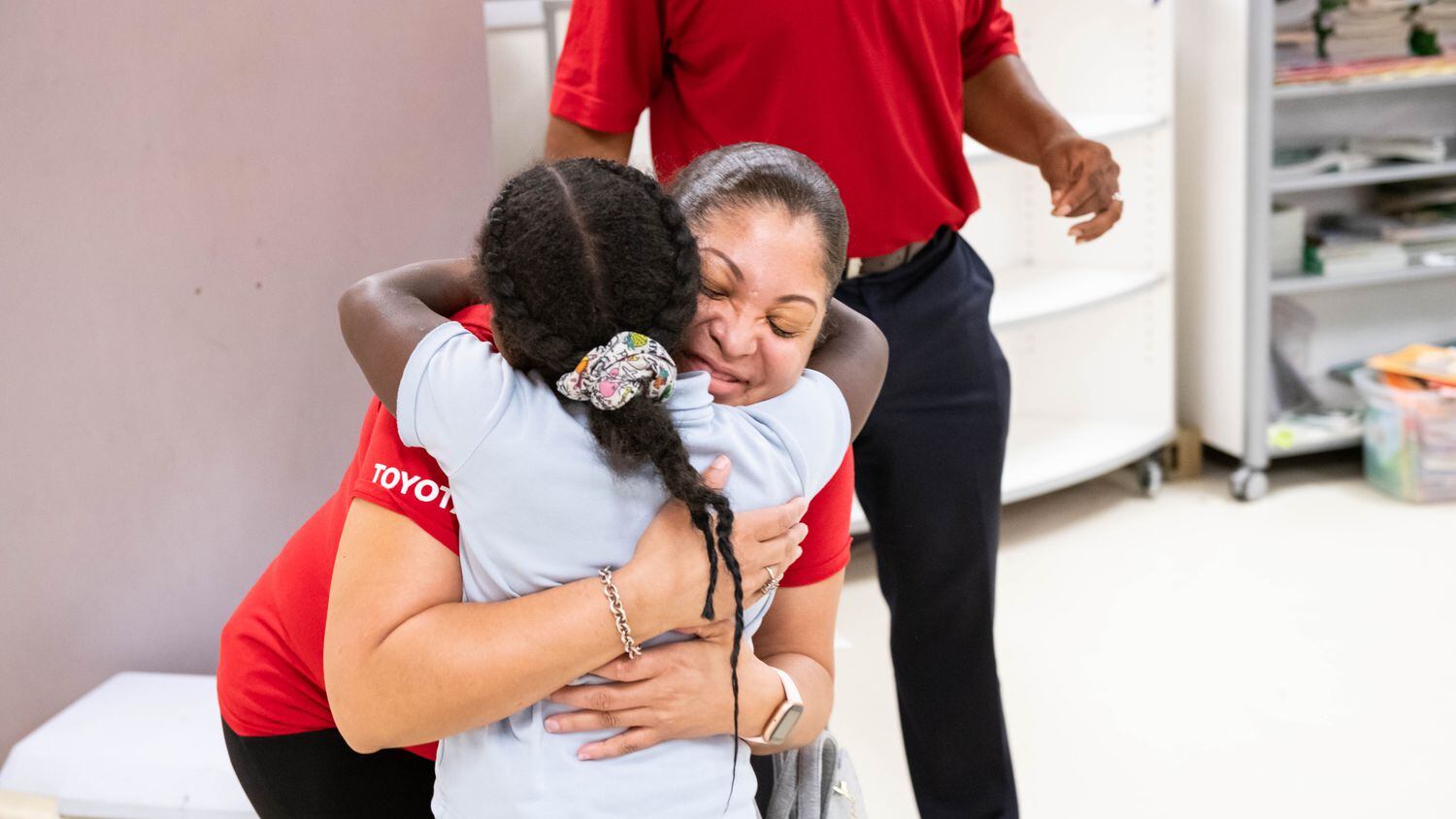 A female volunteer in a red T-shirt hugs a young female student in a classroom.