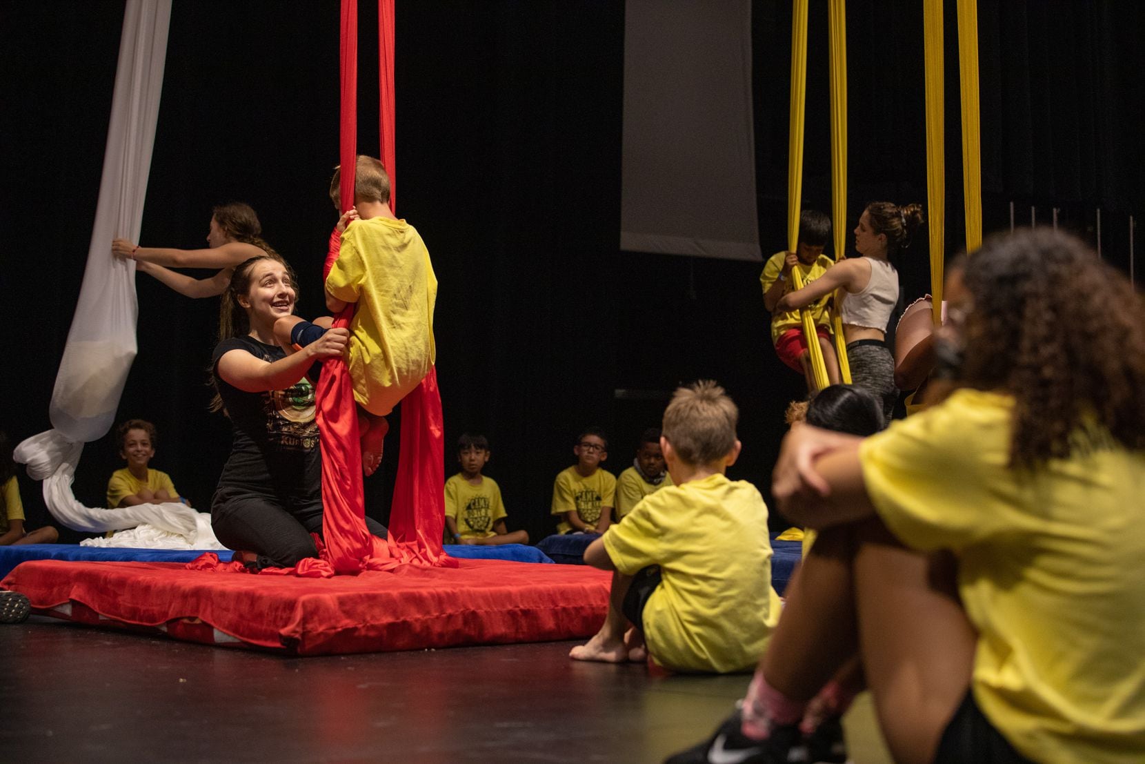 Children in yellow shirts learn to dangle from silk ropes with instructors.
