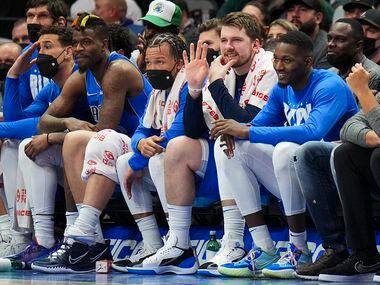 Dallas Mavericks guard Luka Doncic waves to teammates from the bench as he sits between...