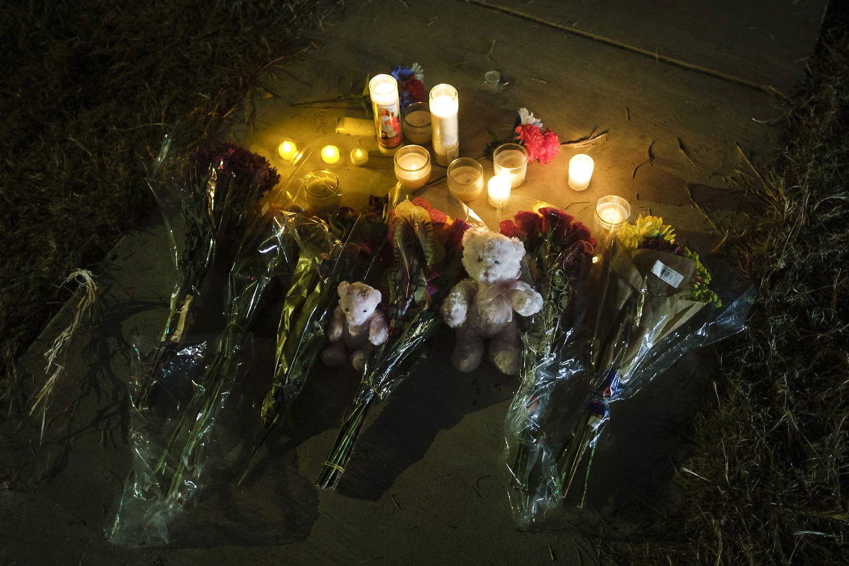 Flowers, candles, and teddy bears rest on the sidewalk in the front yard of the house where...
