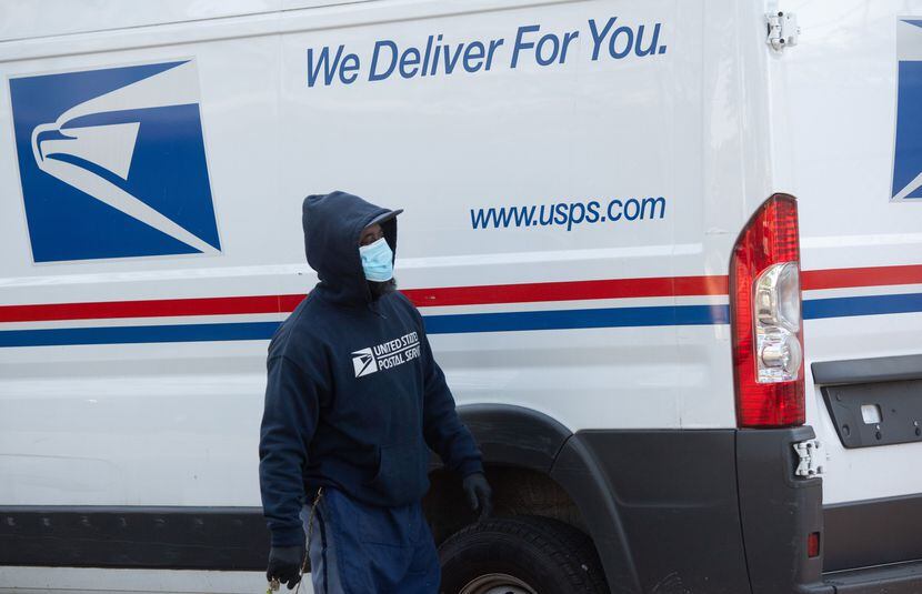 A postal worker wearing a face mask and gloves loads a truck with packages at a post office...