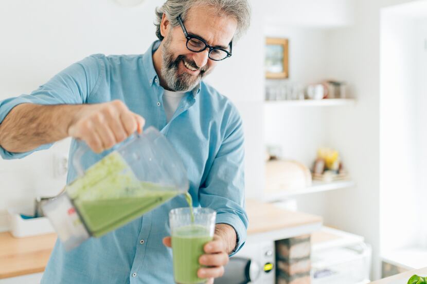A older man pours a green shake from a blender into a glass.