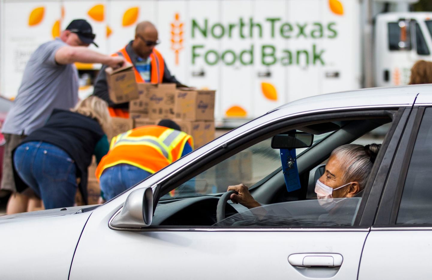 A woman wearing a surgical mask drives past volunteers who prepare to hand out boxes of food...