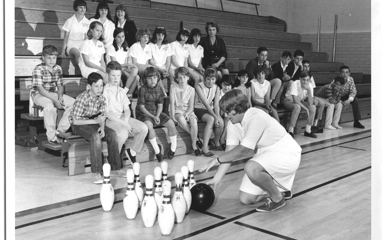 Students sitting in a gymnasium while their gym teacher teaches them how to bowl (Mar. 14,...