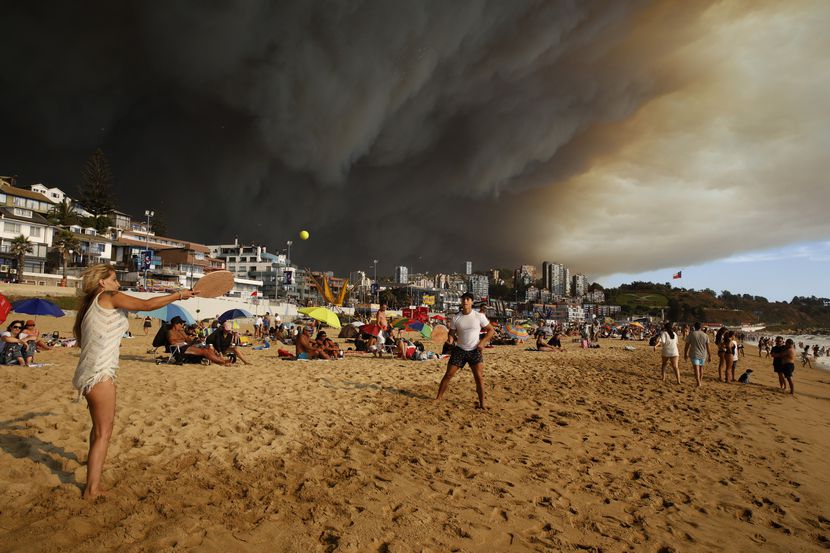 Turistas juegan a las palas en una playa con una gran nube de humo negro a sus espaldas,...