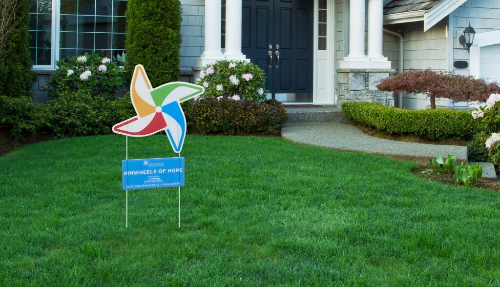 A yard sign in the shape of a pinwheel, in the middle of a green lawn with a house in the...