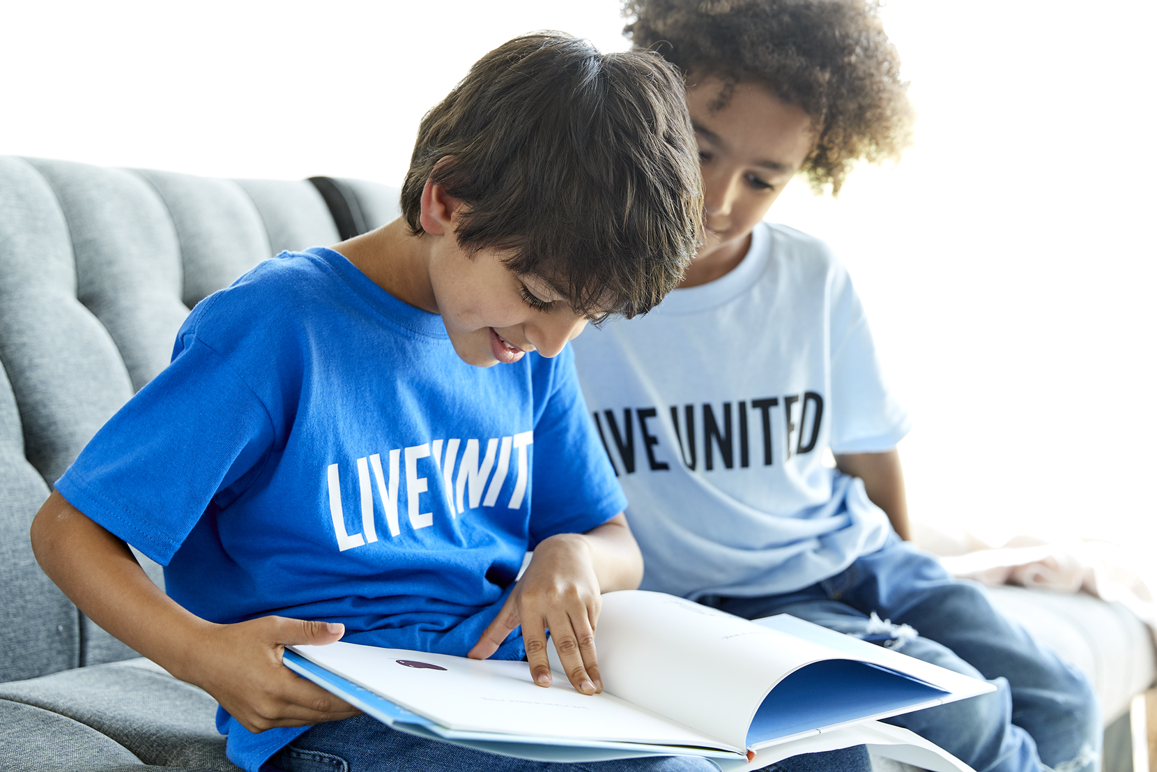 Two young boys read a book while sitting on a couch