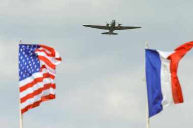 
An American and a French flag are seen in the foreground as a C-47 military transport...