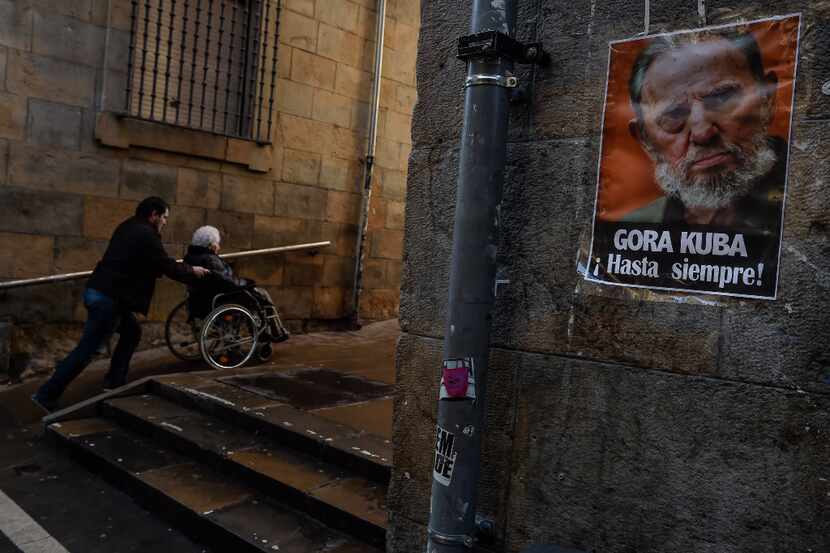 A man wheels a woman in a wheelchair near to a poster with the image of former Cuban...
