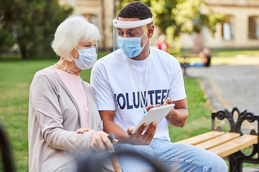 A man in a mask and white volunteer T-shirt helps an elderly woman sitting on park bench.