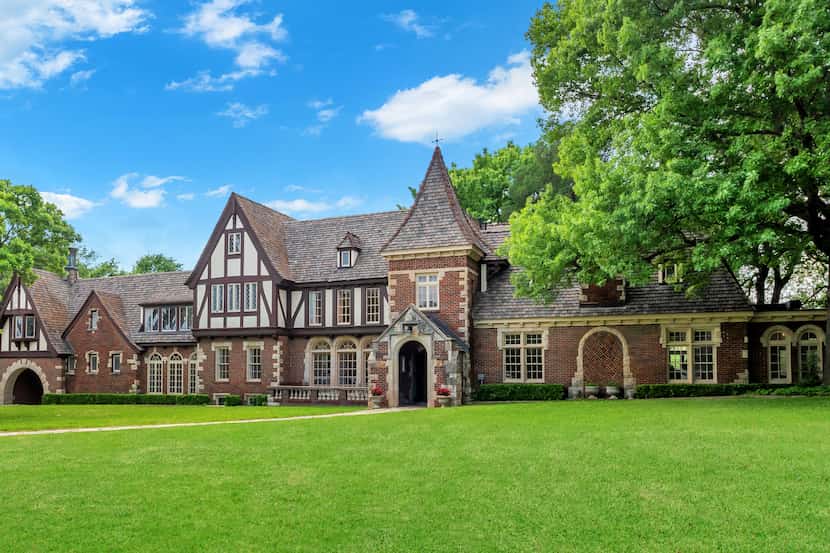 A Norman-Jacobethan Revival home is surrounded by green trees and has a large, vibrant lawn...