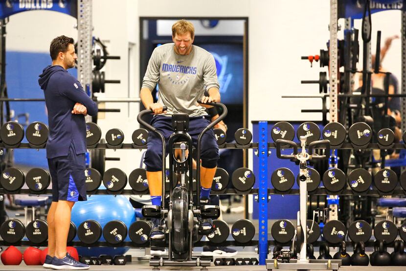 Mavericks forward/center Dirk Nowitzki rides an exercise bike under the watchful eye of...