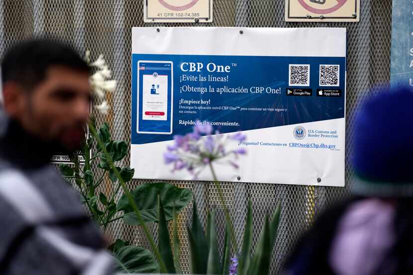 People waiting to apply for asylum stand in front or a sign for the CBP One app as they camp...
