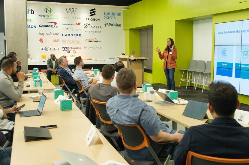 A woman in an orange jacket and jeans speaks to a room of people sitting at conference tables.