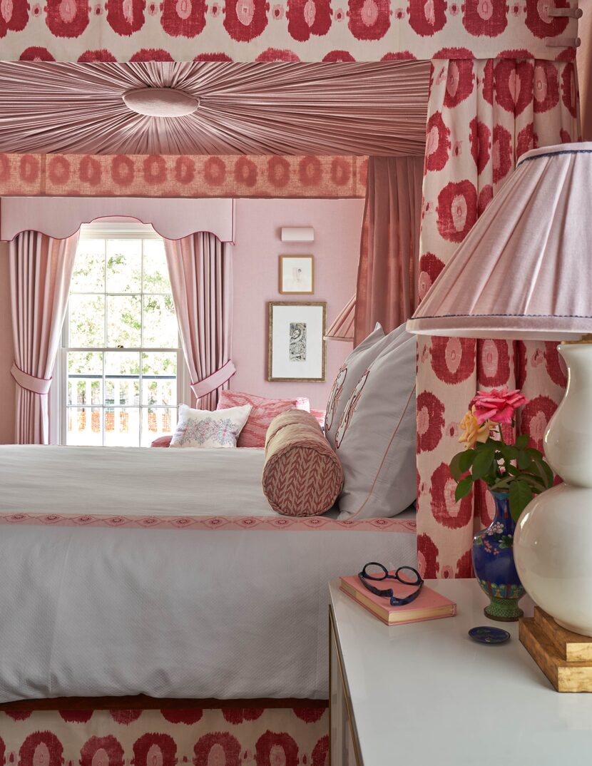 A guest bedroom features a four-poster bed with a printed fabric canopy and pink upholstered...