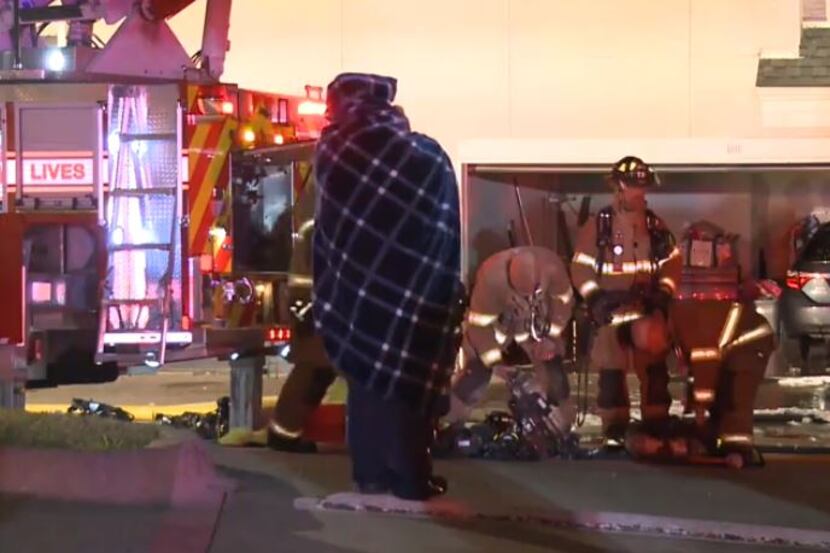 A resident wrapped in a blanket to ward off the cold watches as Plano firefighters battle an...