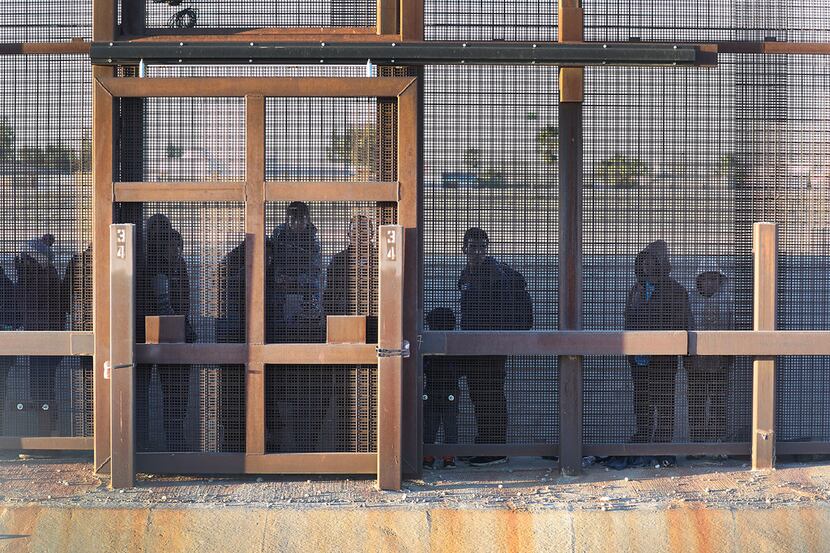 Migrants stand together along the U.S.-Mexican border fence as they wait to turn themselves...