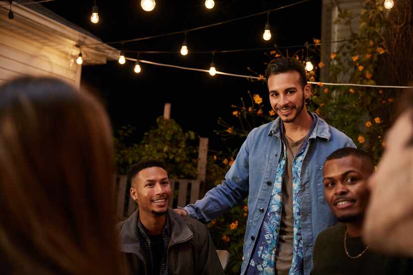 A Latino man stands with a group of friends in a backyard under string lights.