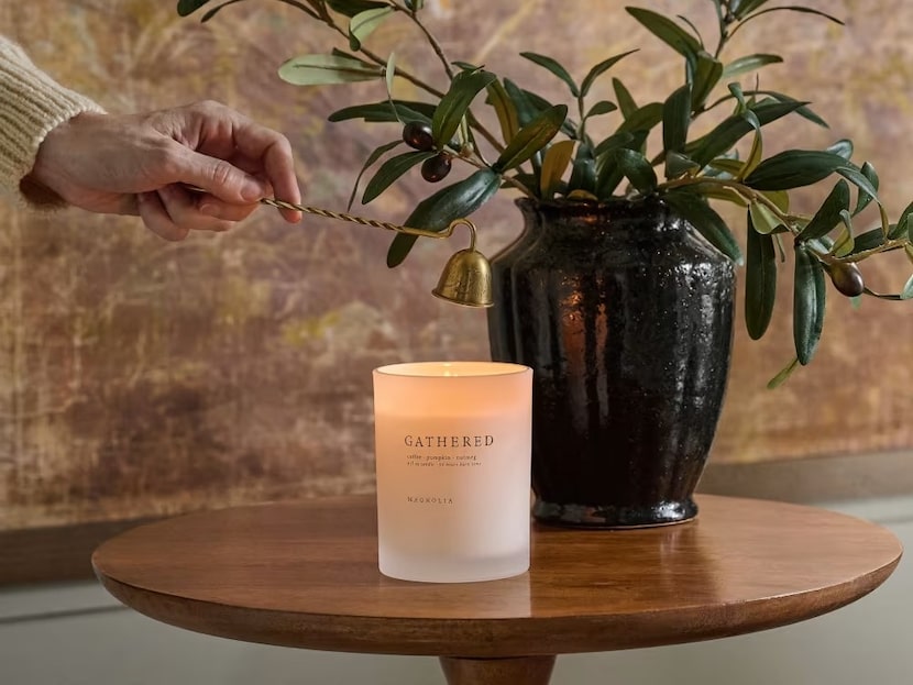 Candle on a table with vase of greenery in the background.