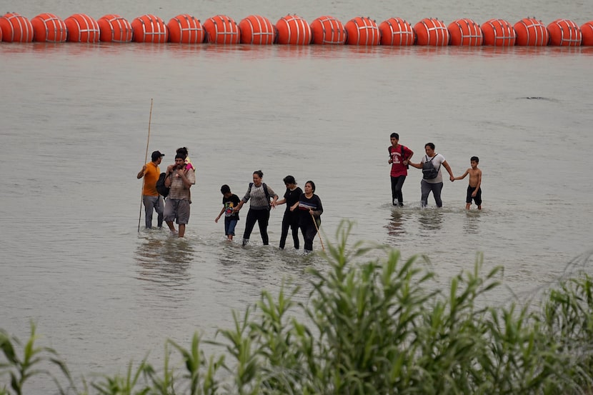 Migrants who crossed the Rio Grande from Mexico walk past a floating barrier near Eagle...