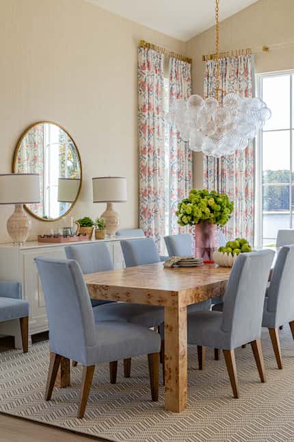 Dining room with blue chairs around a wooden table, atop a patterned rug; colorful drapes...