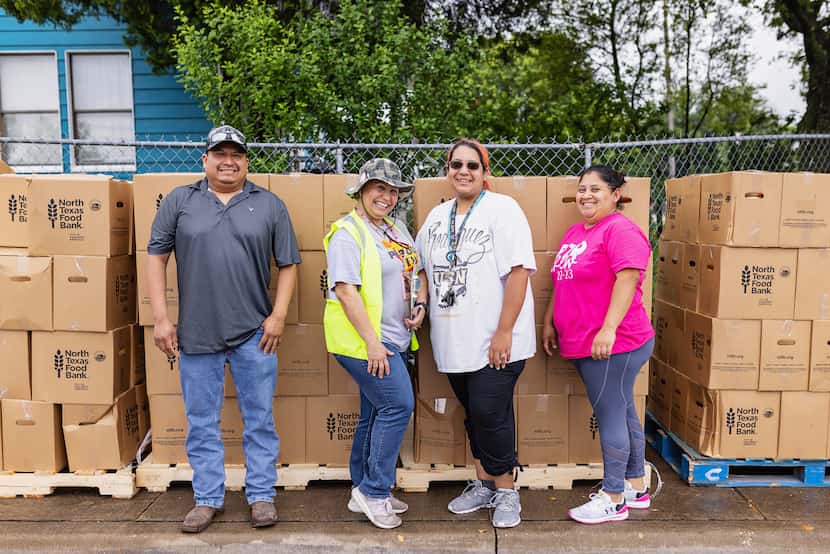 Four volunteers stand in front of pallets stacked with cardboard boxes for food pantry...