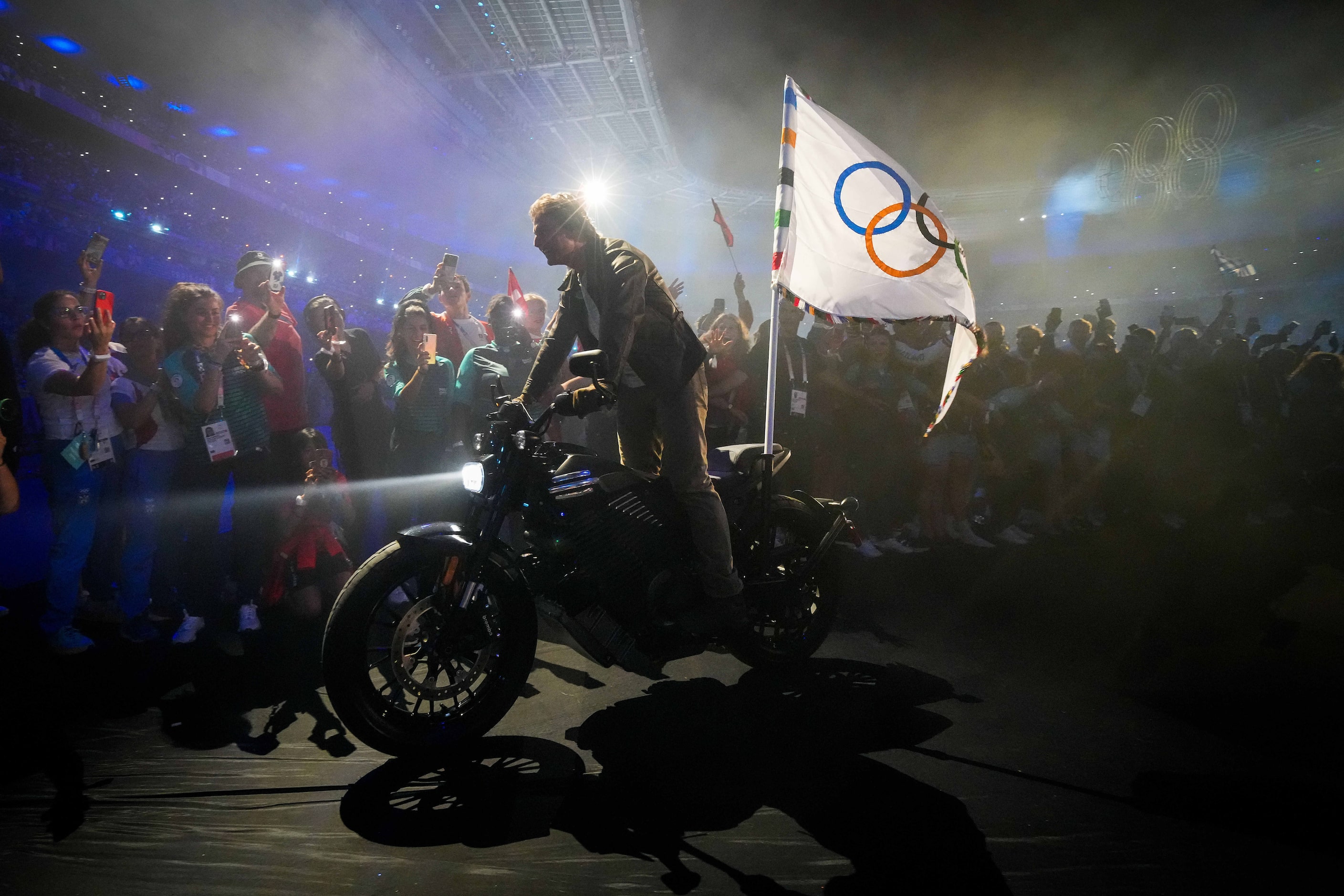 Actor Tom Cruise rides through athletes on a motorcycle carrying the Olympic flag after a...