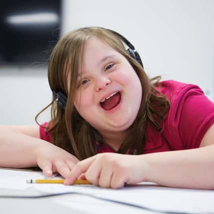 A young girl with Down's Syndrome wears headphones and holds a pencil while she does her...