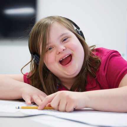 A young girl with Down's Syndrome wears headphones and holds a pencil while she does her...