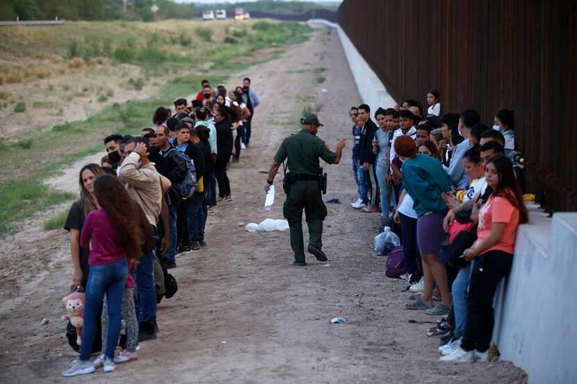 Un grupo de migrantes permanecen junto al muro de la frontera con México mientras un agente...