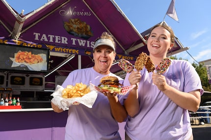 Two women stand in front of a concession stand holding fair confections such as hamburgers...