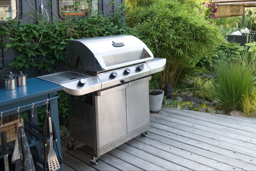 Stainless gas grill with black side, with side table with tools and greenery in the background