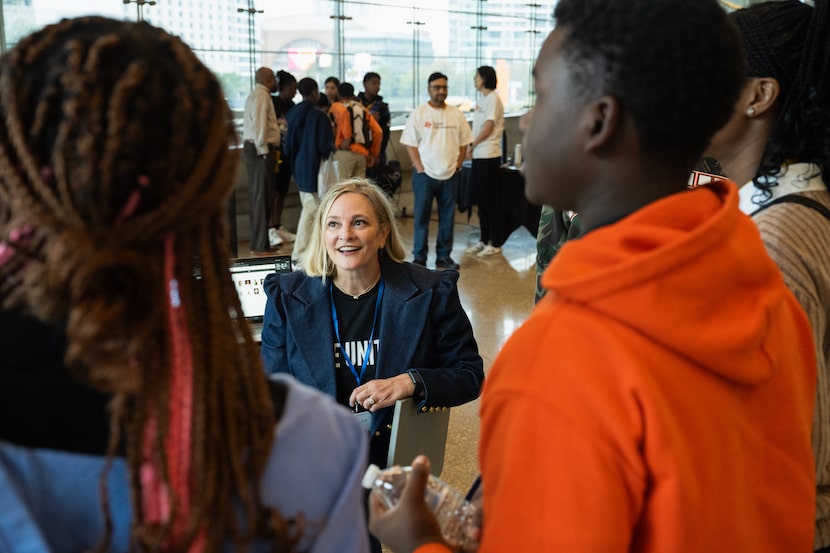 A woman is seated speaking to teenage students who stand in front of her and around her.